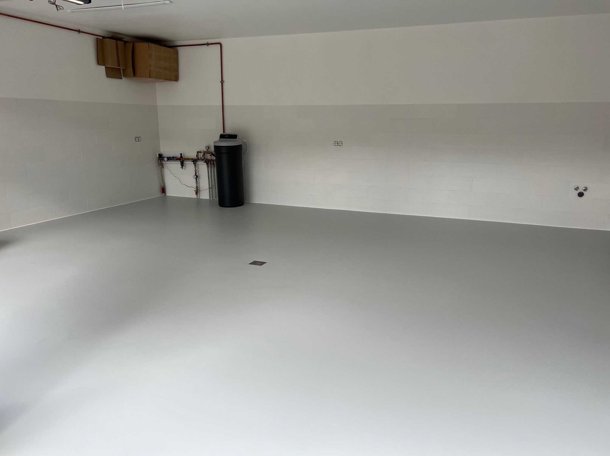 Jasnoszara, gładka posadzka żywiczna w pustym pomieszczeniu z białymi ścianami, widoczne instalacje i czarny zbiornik na ścianie, kartony pod sufitem.