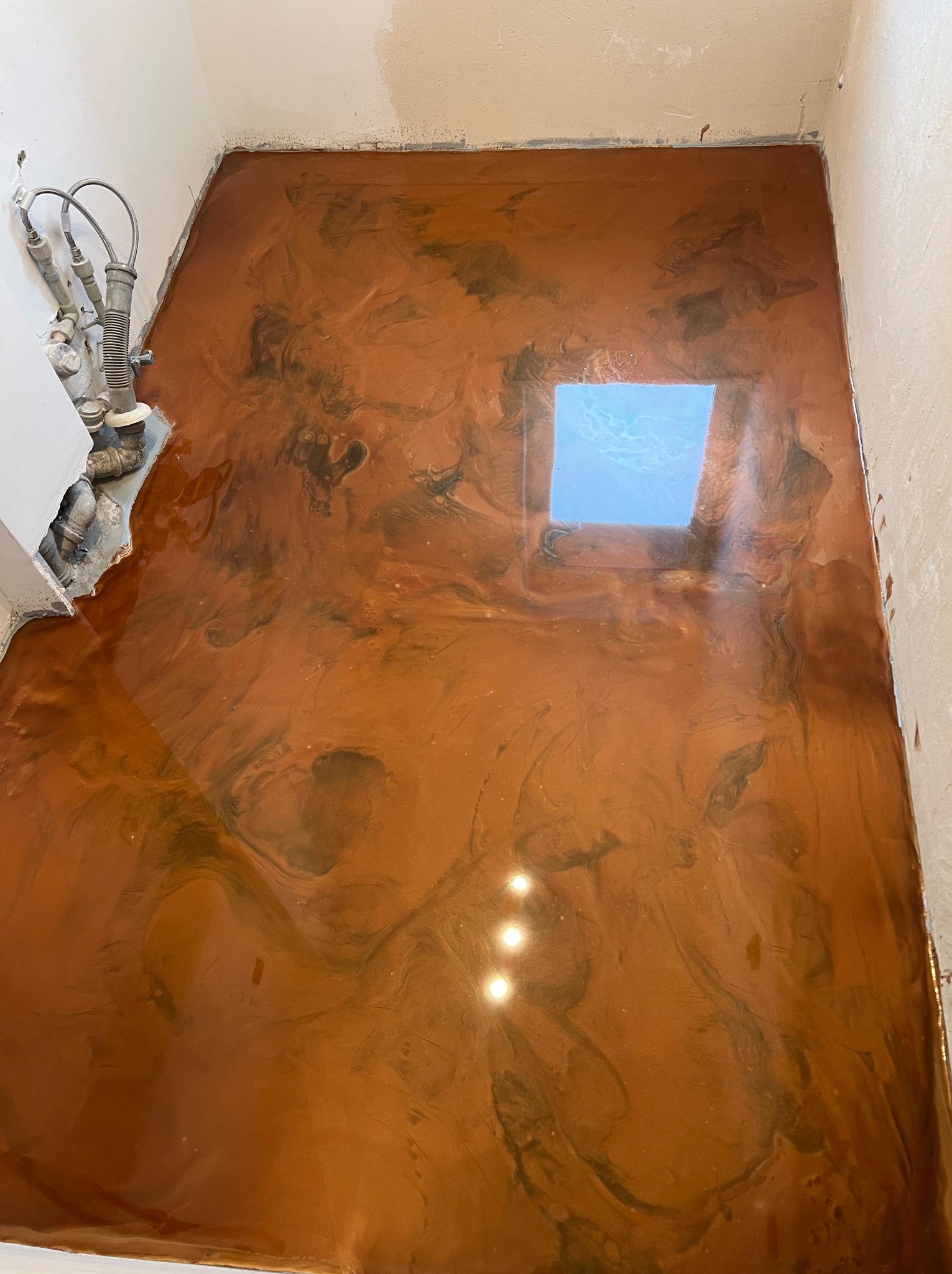 Błyszcząca, brązowa podłoga z efektem marmuru, refleksy świetlne i widoczne rury przy ścianie.