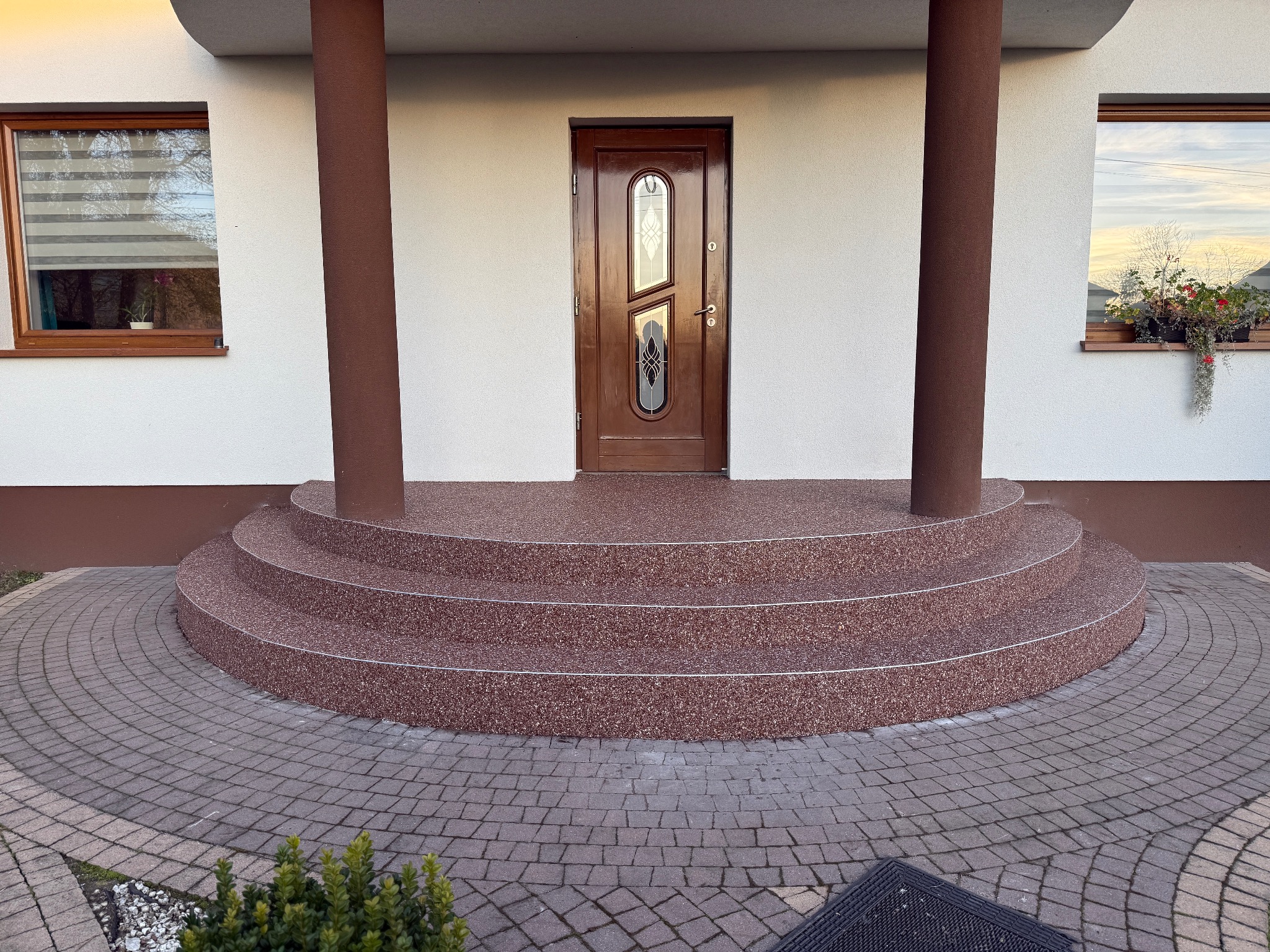 Brązowe schody wejściowe z żywicy, wykończone srebrnymi listwami, prowadzące do drzwi wejściowych. Brukowana ścieżka i kolumny podtrzymujące zadaszenie.