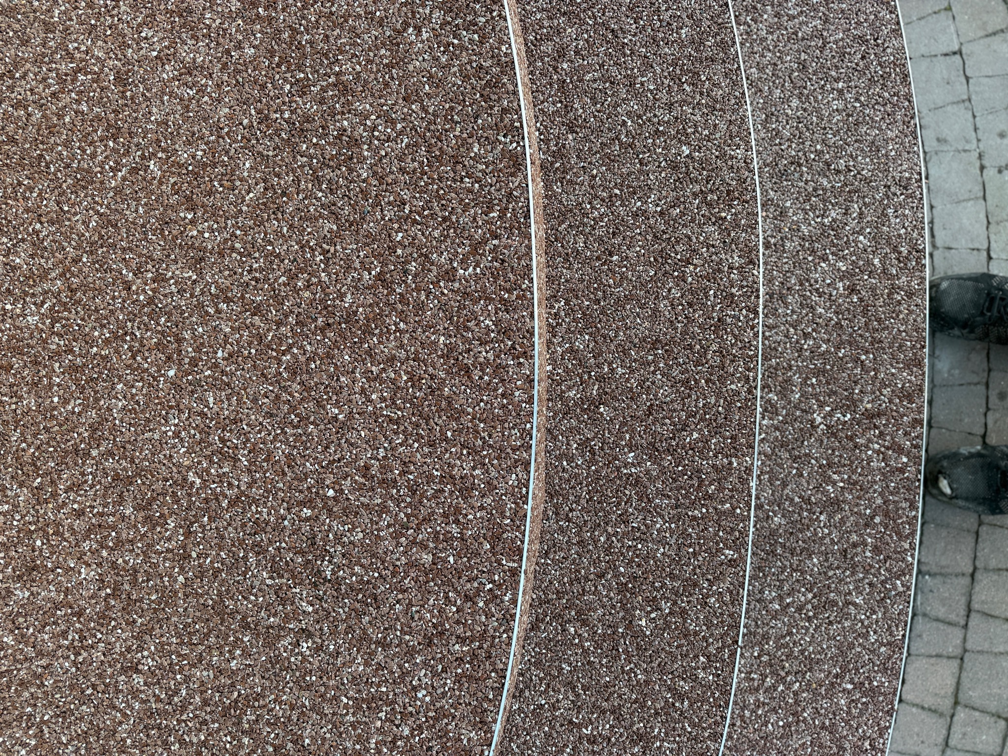 Brązowa, drobnoziarnista powierzchnia z żywicy epoksydowej, widok z góry, z fragmentem buta i kostki brukowej w prawym dolnym rogu. Układ warstwowy.