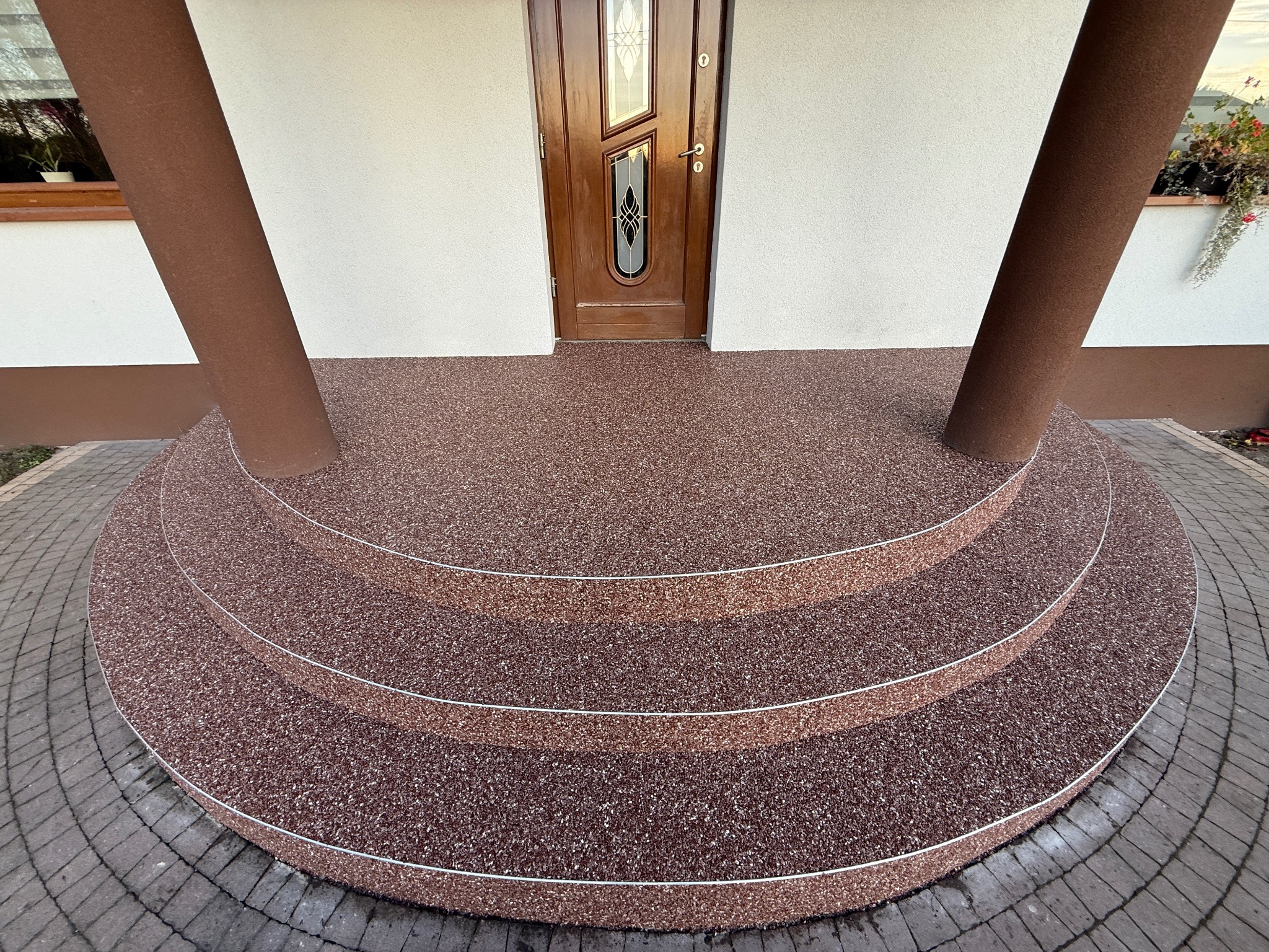 Wykończone schody wejściowe z brązową, kamyczkową nawierzchnią, otoczone szarą kostką brukową. Widoczne brązowe kolumny i drewniane drzwi wejściowe.