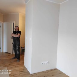 Usługi Remontowe - Świeżo pomalowane na jasny beż narożne ściany w pokoju z drewnianą podłogą, widoczny elektryk w kombinezonie z założonymi rękami i puszka farby w rogu pomieszczenia.