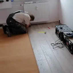 Pracownik w kombinezonie montuje panele podłogowe w pokoju z grzejnikiem, widoczna podkładka korkowa i narzędzia w skrzynkach.