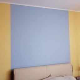 Usługi Remontowe - Ściana w pokoju pomalowana na dwa kolory: żółty i niebieski, z widocznym fragmentem łóżka z beżowym zagłówkiem i lampką nocną.