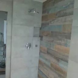 Prysznic z mieszanką płytek: szare, betonowe płytki na ścianie z armaturą i deszczownicą oraz dekoracyjna ściana z płytek imitujących drewno w odcieniach brązu, szarości i błękitu.