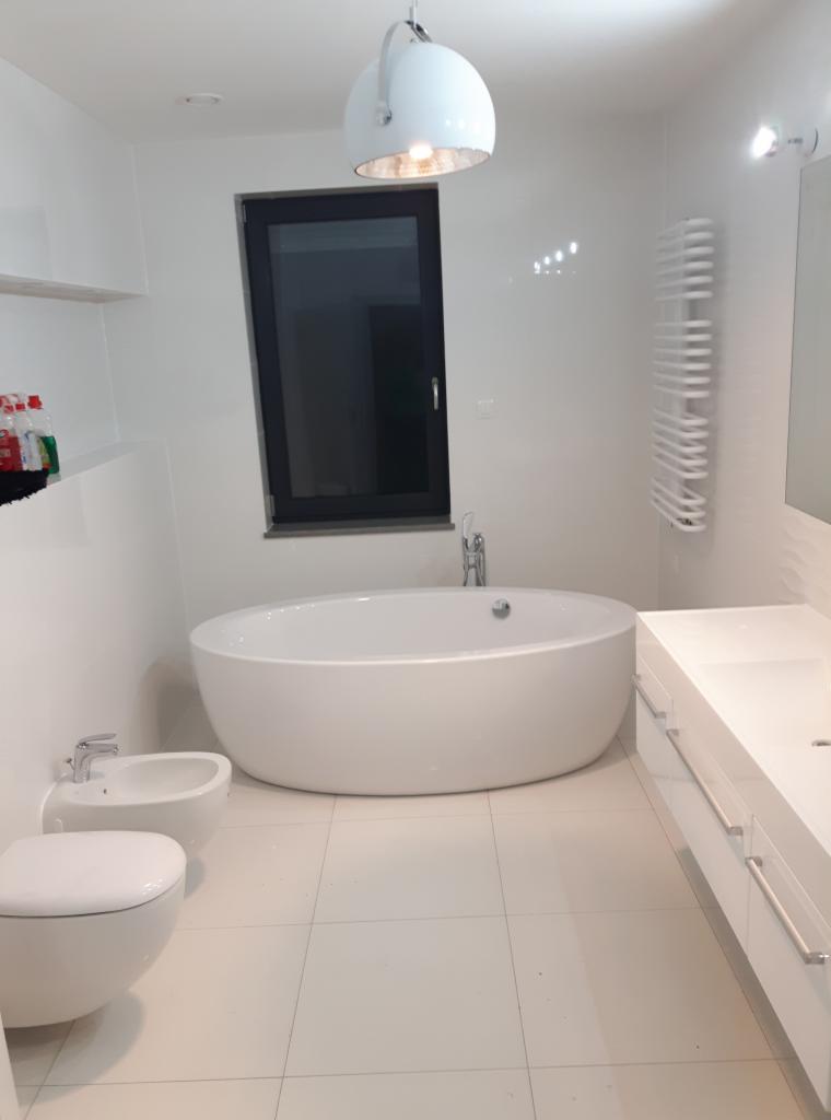 Nowoczesna, minimalistyczna łazienka w bieli z wolnostojącą wanną, bidetem, podwieszaną toaletą i grzejnikiem drabinkowym na ścianie, oświetlona lampą wiszącą w kształcie kuli.