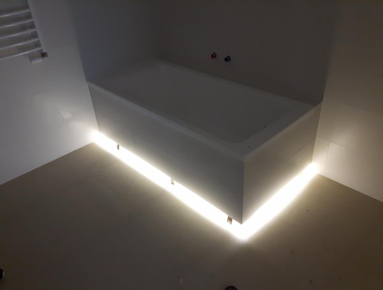 Biała wanna z podświetleniem LED u dołu, zamontowana w łazience z jasnymi płytkami i grzejnikiem drabinkowym na ścianie.