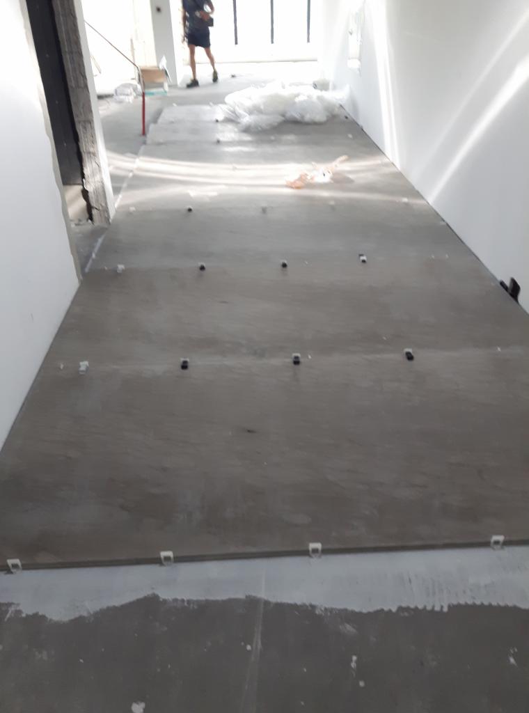 Wyrównana, szara podłoga z widocznymi czarnymi i białymi elementami poziomującymi, w tle sylwetka osoby i materiały budowlane