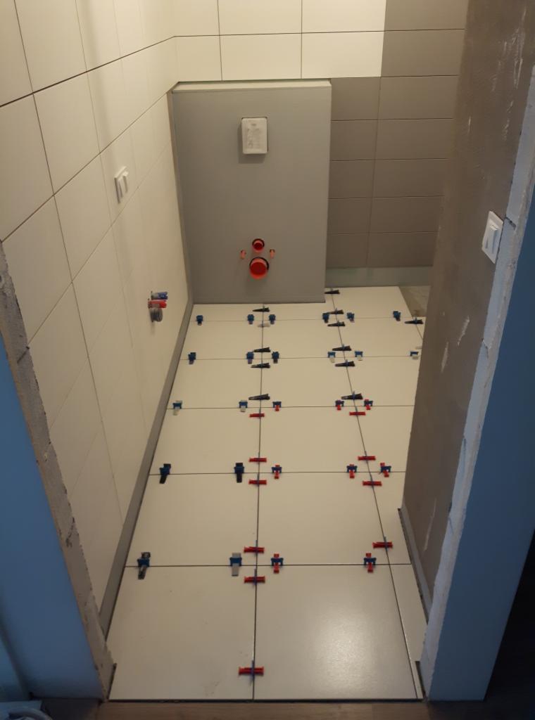 Białe płytki podłogowe w trakcie układania w łazience, z użyciem czerwono-niebieskich klinów poziomujących, widoczne pionowe rury kanalizacyjne w ścianie.