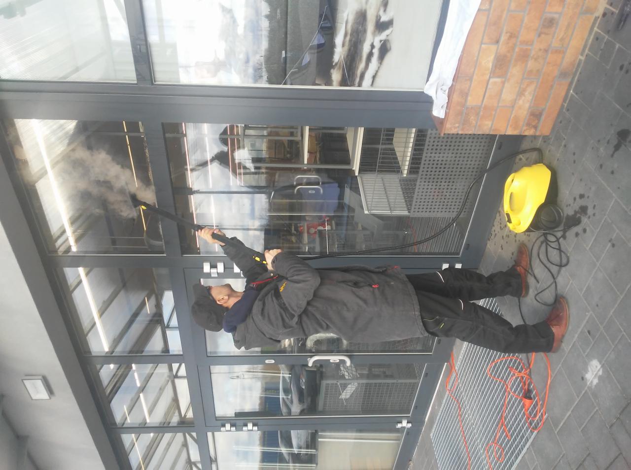 Pracownik w kurtce i czapce czyści parą okna na zewnątrz budynku, używając żółtego parownika Kärcher; widoczne odbicie w oknach, szare ramy okienne i brązowe buty pracownika.