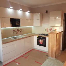 Jasna kuchnia z drewnianym blatem, kwiatowym motywem na szklanym panelu nad blatem, widoczne kartony zabezpieczające podłogę podczas montażu.