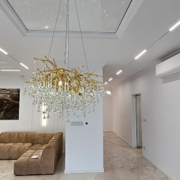 Wnuk Budownictwo - Nowoczesny salon z beżową sofą i błyszczącą podłogą z płytek imitujących marmur. W centrum złoty żyrandol z kryształkami, a na suficie oświetlenie punktowe.