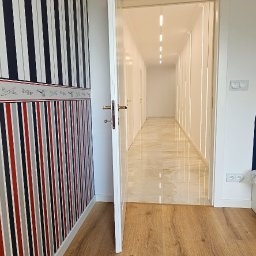 Wnuk Budownictwo - Wnętrze korytarza z wzorzystą tapetą w paski i drewnianą podłogą przechodzącą w marmurową posadzkę. Otwarta biała  futryna drzwiowa. Oświetlenie liniowe.