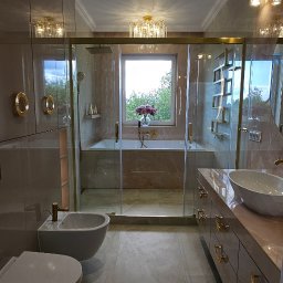 Wnuk Budownictwo - Elegancka łazienka w beżach z marmurkową teksturą, kabina prysznicowa ze złotymi detalami, bidet i umywalka na szafce. Widok na okno z zielenią.