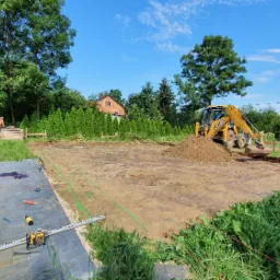 Przygotowanie terenu pod budowę domu: żółta koparko-ładowarka JCB wyrównuje ziemię, widoczne wytyczone granice fundamentów, narzędzia pomiarowe i plan budowy.