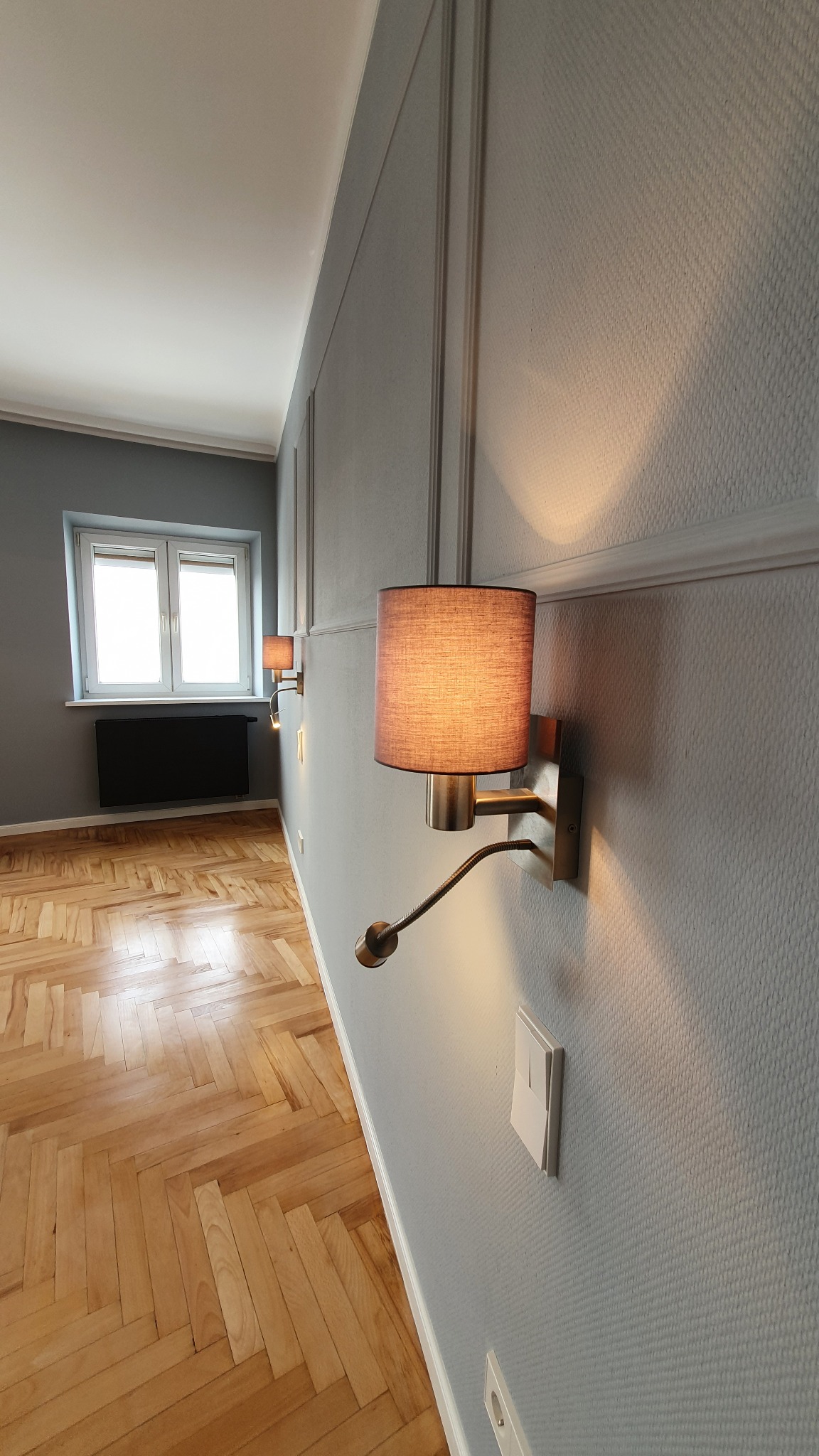 Szary pokój po remoncie: parkiet w jodełkę, ściany z dekoracyjnymi listwami i tapetą, dwa kinkiety z abażurami, okno z grzejnikiem. Nowoczesne wykończenie wnętrza.