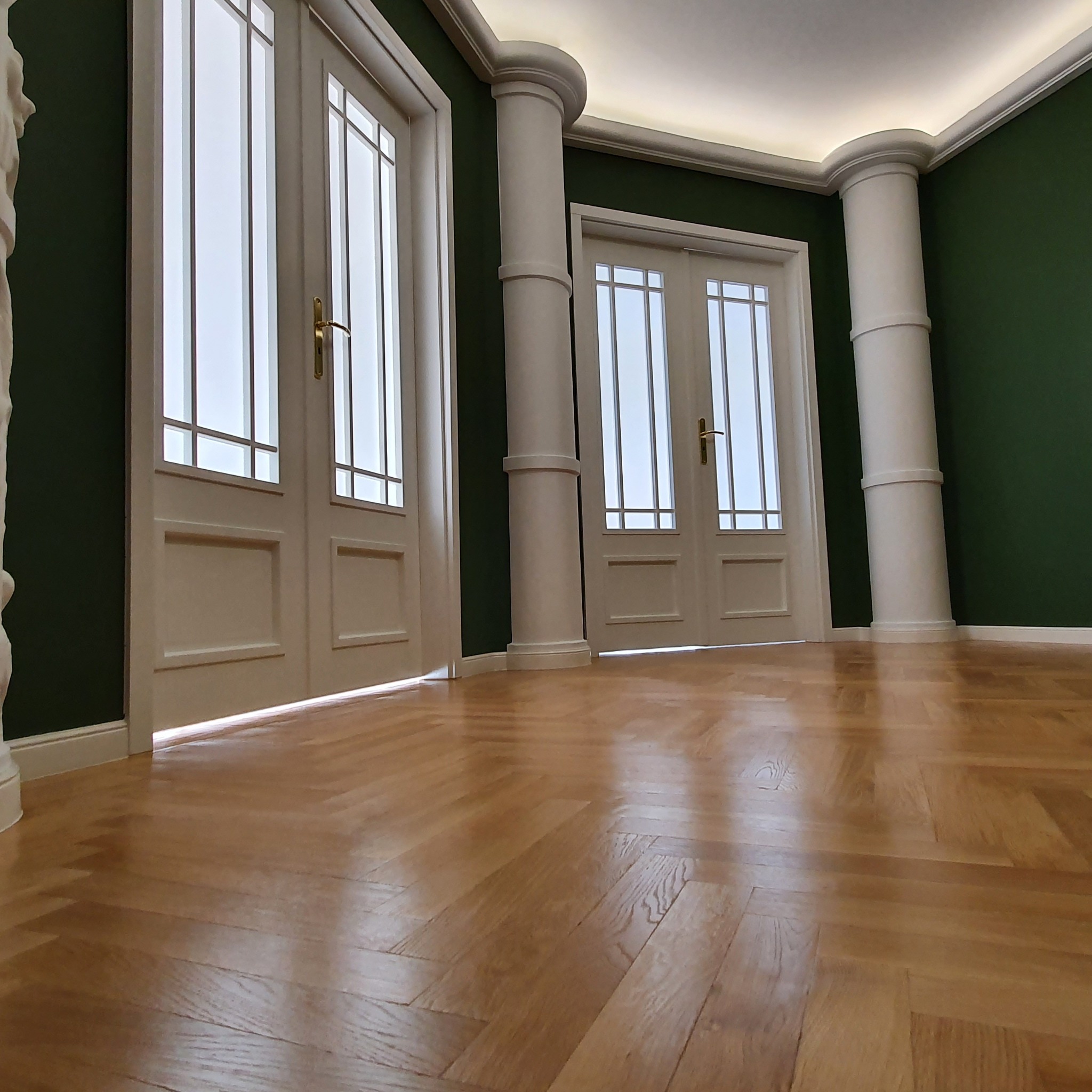 Eleganckie wnętrze z parkietem w jodełkę, białymi kolumnami i stolarką okienną. Ściany w odcieniach butelkowej zieleni. Widok z niskiej perspektywy.