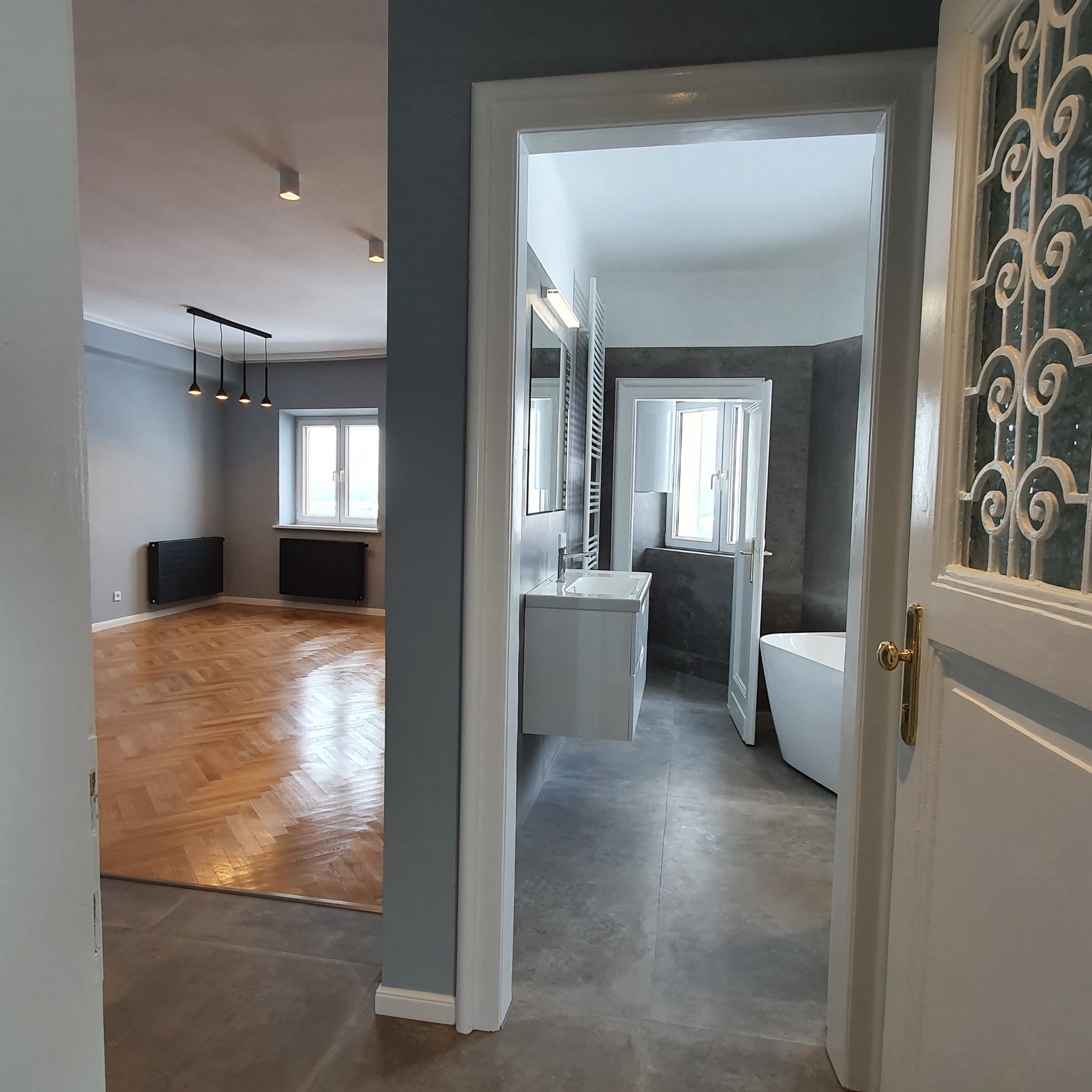 Widok przez otwarte drzwi na odnowioną łazienkę z białą wanną i umywalką oraz na pokój z parkietem i grzejnikami. Szara kolorystyka ścian.