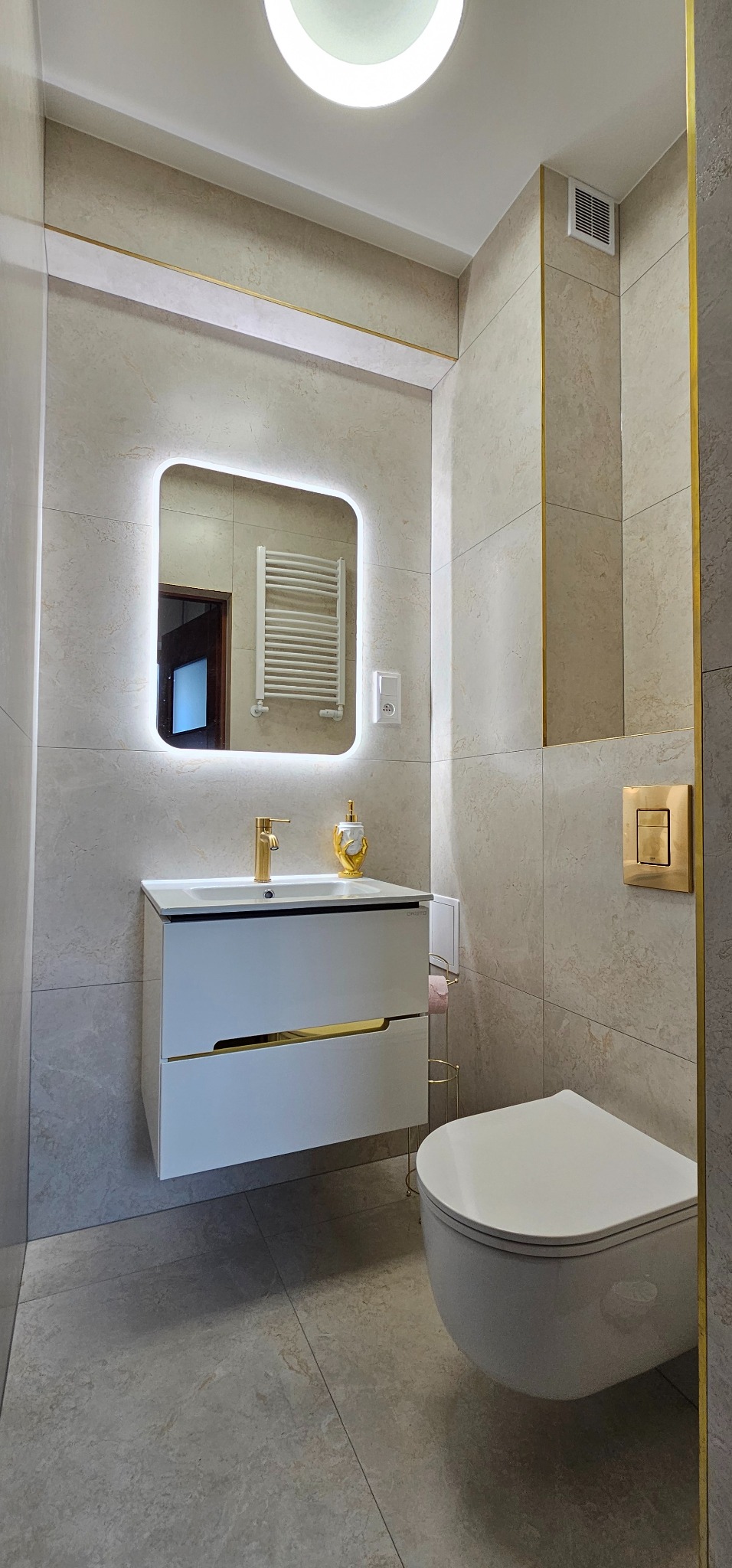 Nowoczesna łazienka w beżowych płytkach z złotymi akcentami. Umywalka z szafką, podświetlane lustro i wisząca toaleta. Widoczny fragment grzejnika w odbiciu lustra.