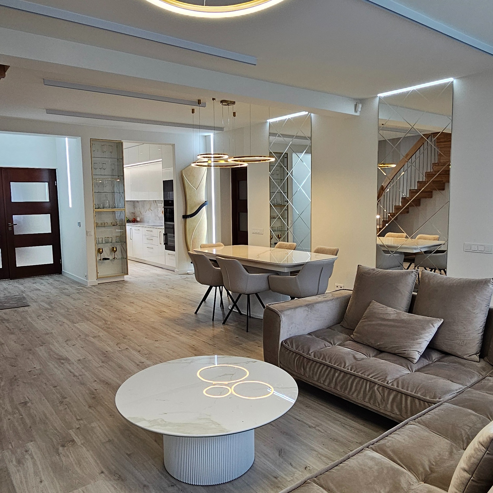 Nowoczesne wnętrze salonu z jadalnią: beżowa sofa, okrągły stolik z podświetleniem, stół z krzesłami, lustra na ścianie i oświetlenie LED. Parkiet w odcieniach szarości.