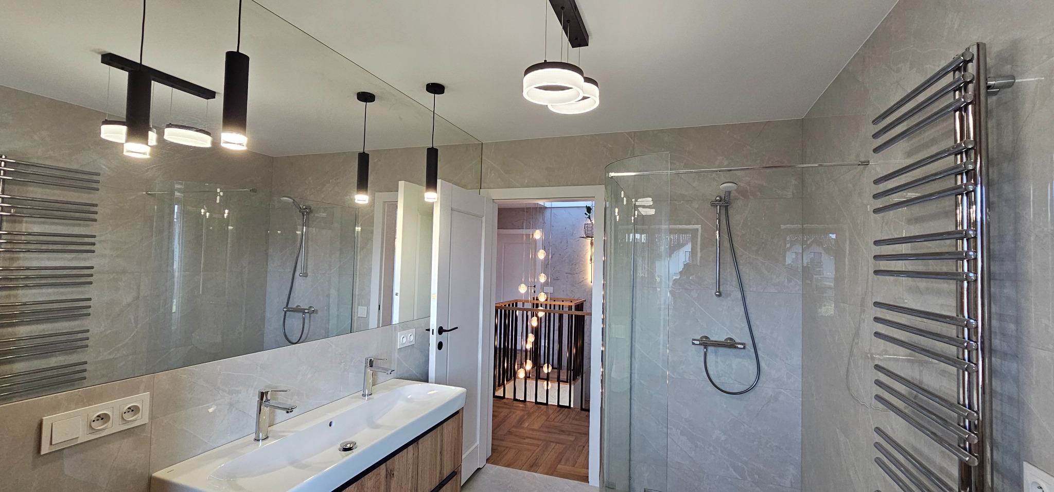 Nowoczesna łazienka z kabiną prysznicową, umywalką na szafce z drewnianym frontem i grzejnikiem drabinkowym. Widoczne oświetlenie sufitowe i lustro.