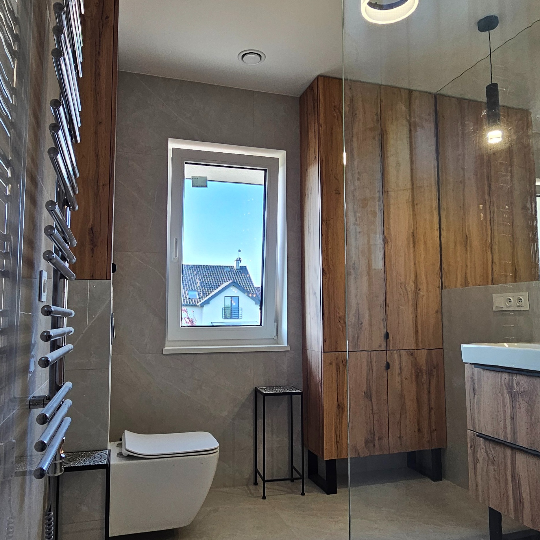 Nowoczesna łazienka z drewnianymi szafkami, grzejnikiem drabinkowym i oknem z widokiem na dom. Uwagę zwraca mały, czarny stolik obok toalety.