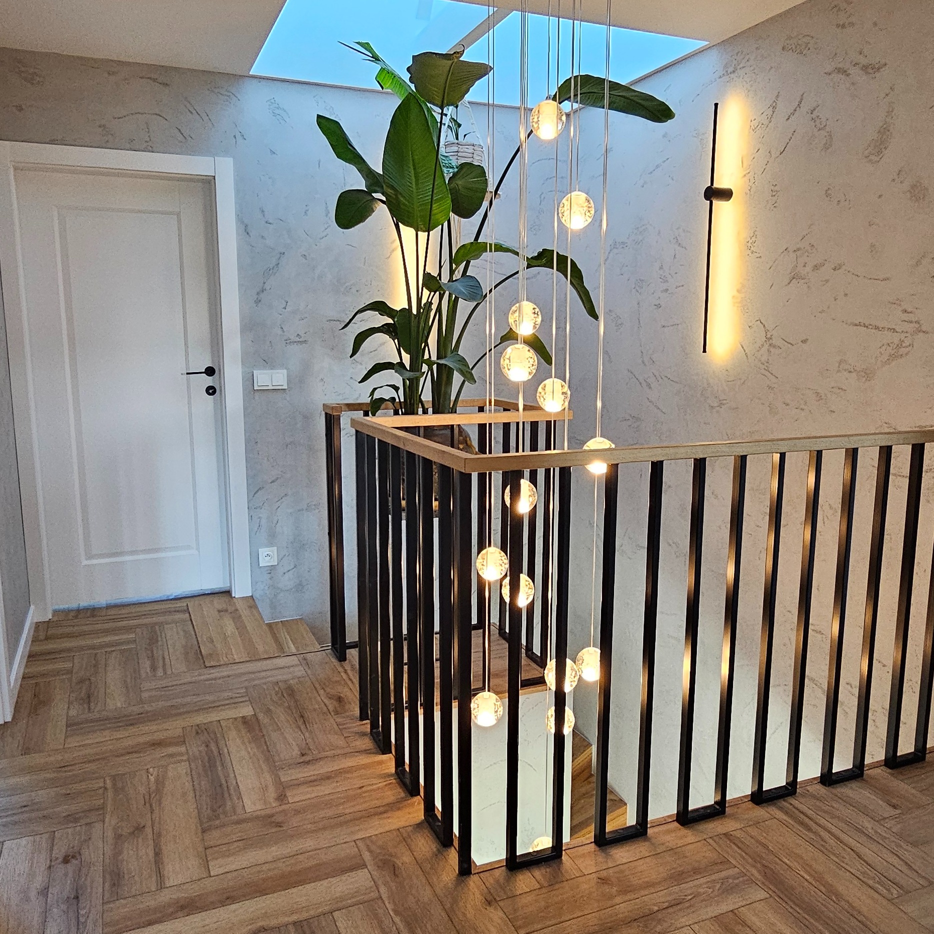 Stylowe wykończenie wnętrza: drewniana podłoga w jodełkę, balustrada z czarnych prętów, roślina doniczkowa i lampa wisząca z kulami w nowoczesnym domu z oknem dachowym.