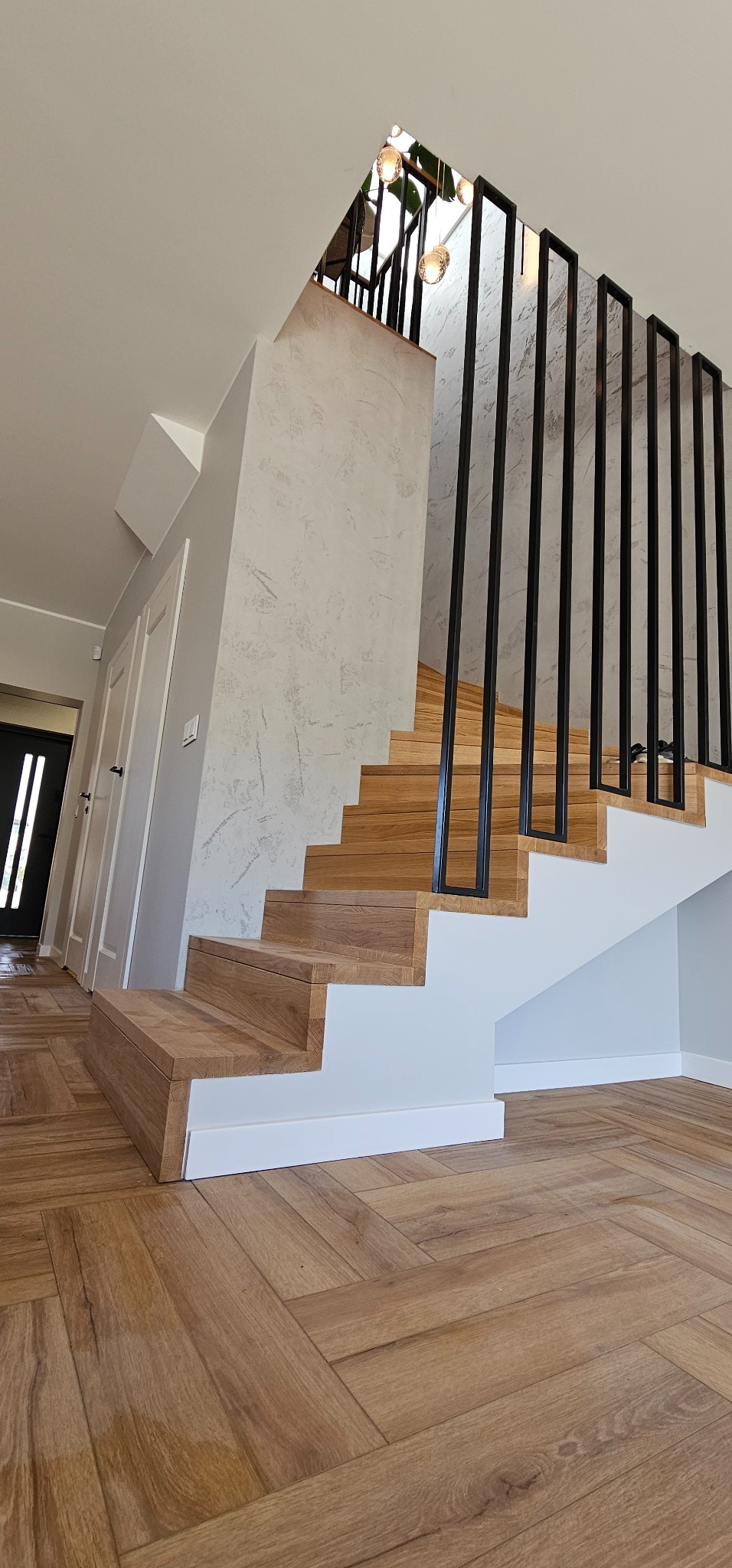 Nowoczesne schody drewniane z czarną balustradą i podświetleniem, wykończone w jasnych barwach, z parkietem ułożonym w jodełkę. Widok z dołu, podkreślający geometrię.