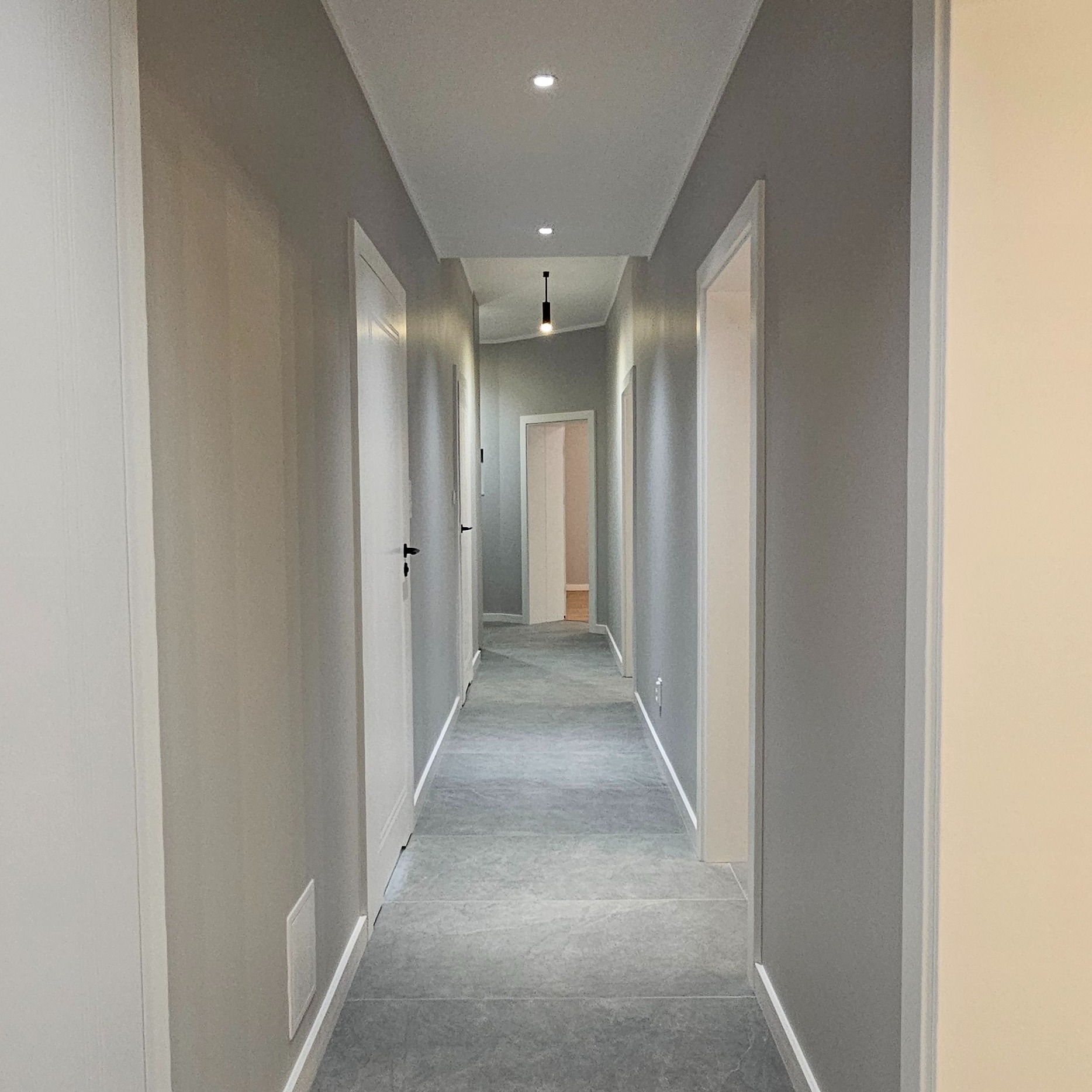 Wykończony korytarz w odcieniach szarości z białymi drzwiami i listwami przypodłogowymi. Widoczne punktowe oświetlenie sufitowe i podłużna lampa. Nowoczesny design.
