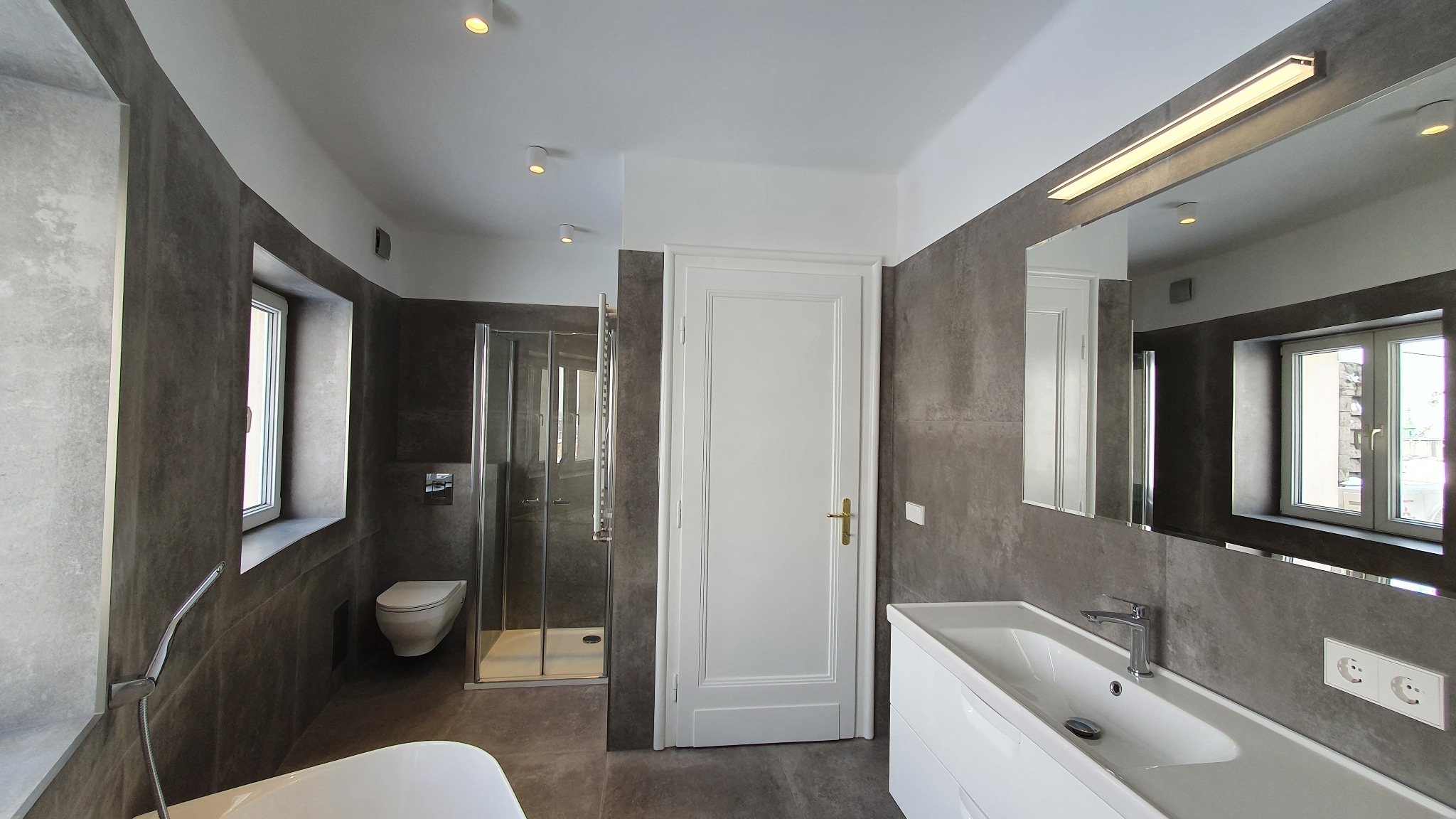 Nowoczesna łazienka z szarymi płytkami imitującymi beton, białą armaturą i kabiną prysznicową. Minimalistyczny design z oświetleniem punktowym i dużym lustrem.