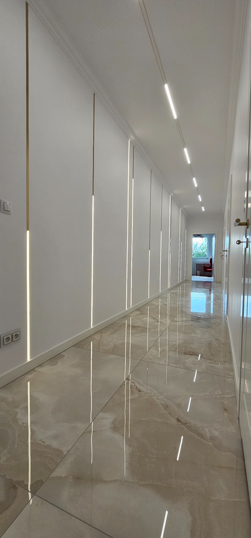 Długi korytarz z błyszczącą podłogą z płytek imitujących marmur. Na ścianach pionowe, podświetlane listwy dekoracyjne i oświetlenie sufitowe.