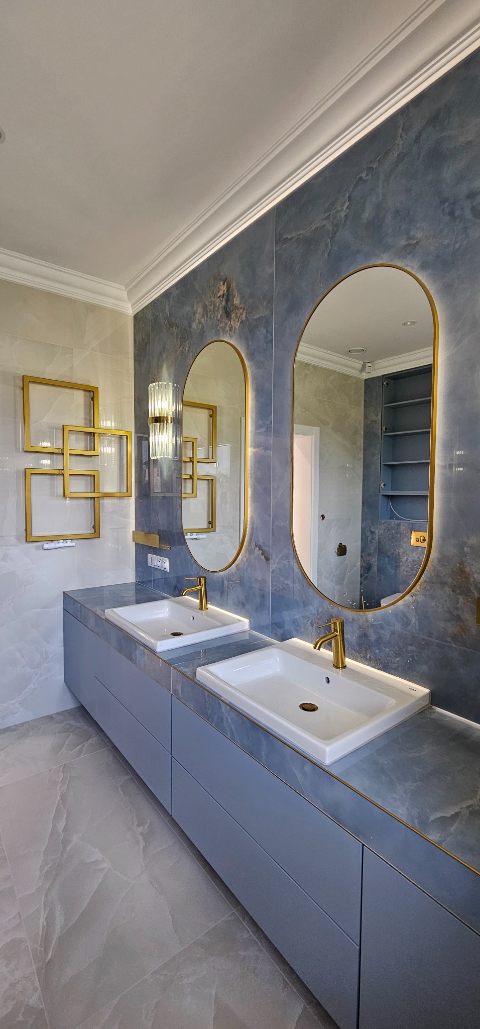 Stylowa łazienka z dwoma umywalkami, lustrami w złotych ramach i marmurowymi płytkami. Uwagę przykuwają geometryczne, złote dekoracje ścienne i oświetlenie.