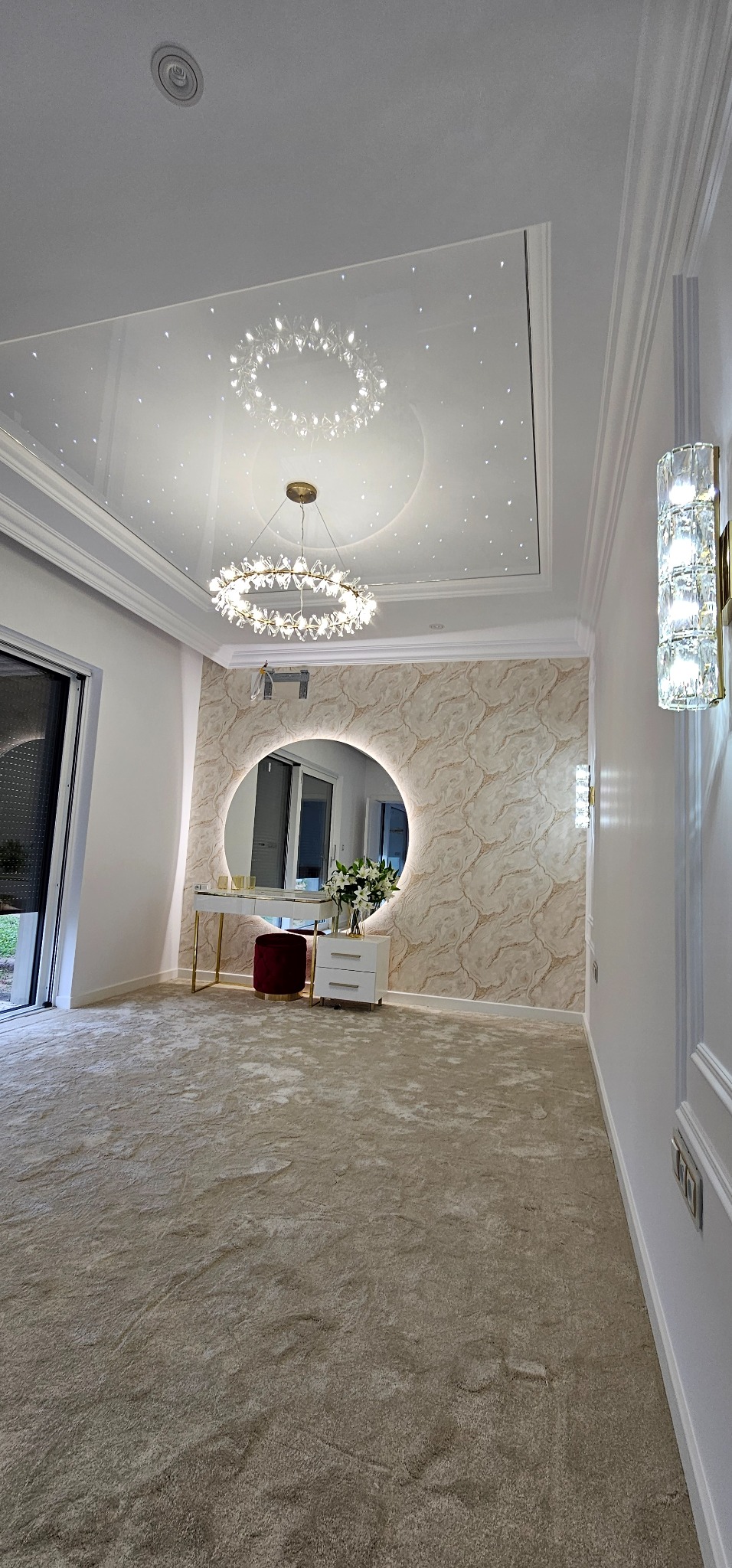 Eleganckie wykończenie wnętrza: okrągłe lustro z podświetleniem, dekoracyjna tapeta, kryształowy żyrandol i lampy ścienne. Beżowy dywan dodaje przytulności.