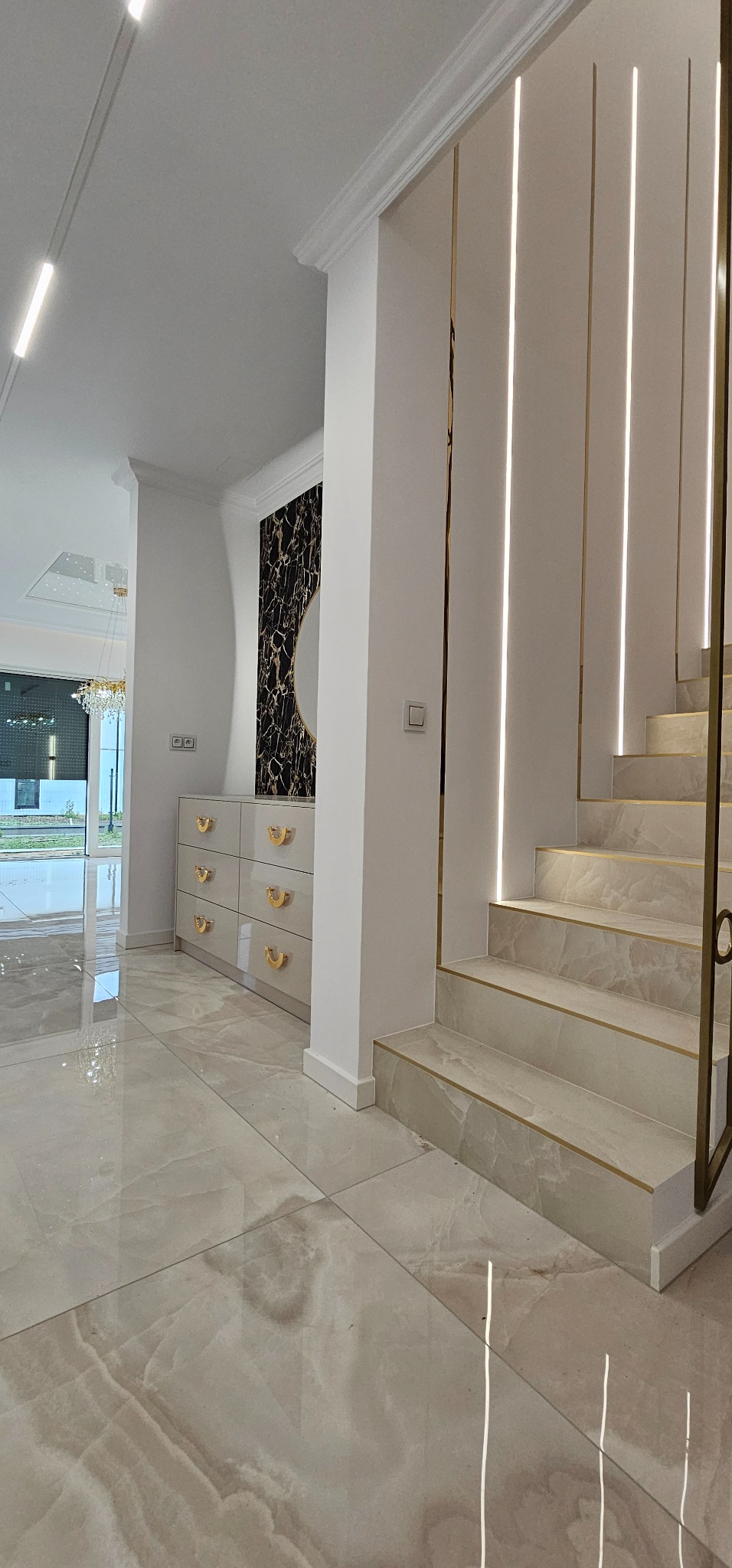 Nowoczesne wnętrze domu: marmurowe płytki podłogowe, komoda z złotymi uchwytami, schody z oświetleniem LED i dekoracyjna ściana z lustrem w złotej ramie.