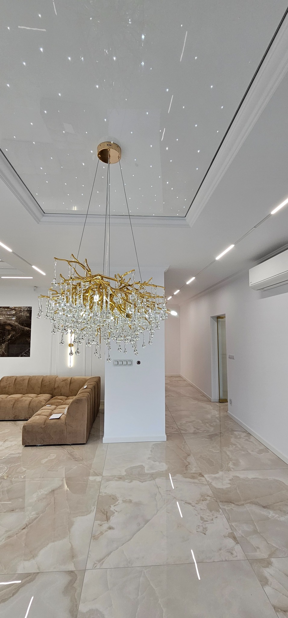 Nowoczesny salon z beżową sofą i błyszczącą podłogą z płytek imitujących marmur. W centrum złoty żyrandol z kryształkami, a na suficie oświetlenie punktowe.