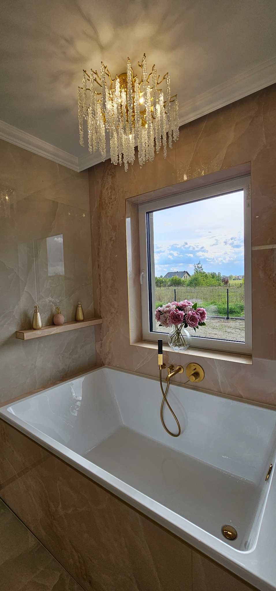 Elegancka łazienka z wanną, marmurowymi płytkami i złotą armaturą. Okno z widokiem na zieleń. Kryształowy żyrandol dodaje luksusowego charakteru.
