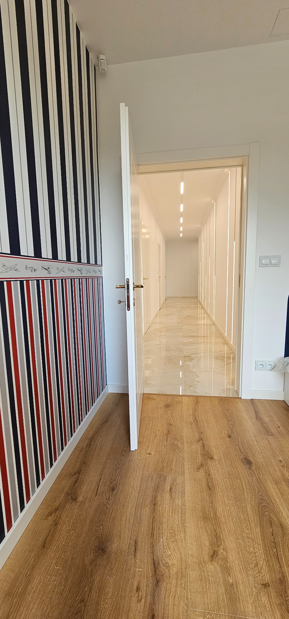 Wnętrze korytarza z wzorzystą tapetą w paski i drewnianą podłogą przechodzącą w marmurową posadzkę. Otwarta biała  futryna drzwiowa. Oświetlenie liniowe.