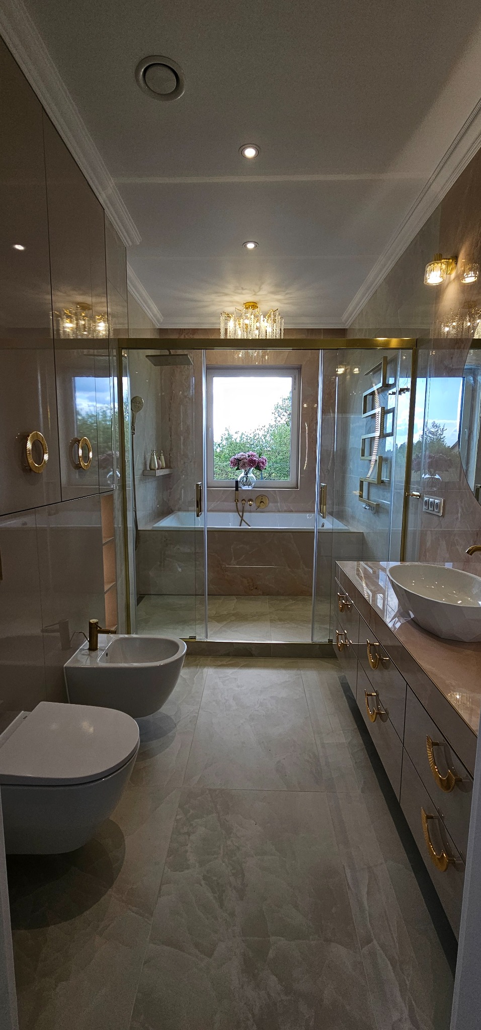 Elegancka łazienka w beżach z marmurkową teksturą, kabina prysznicowa ze złotymi detalami, bidet i umywalka na szafce. Widok na okno z zielenią.