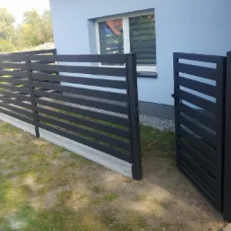 Nowoczesne, poziome ogrodzenie panelowe w kolorze antracytowym z betonową podmurówką i furtką, na tle jasnoniebieskiego domu z białymi ramami okiennymi.