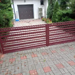 Ciemnoczerwona brama wjazdowa z poziomych profili, osadzona na ceglanych słupkach, na tle kostki brukowej i domu z garażem.