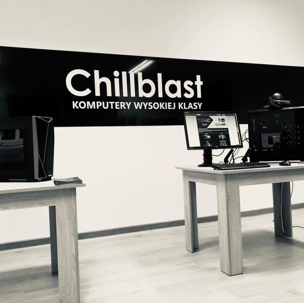 Dwa stanowiska komputerowe z logo 'Chillblast Komputery Wysokiej Klasy' na ścianie, pokazujące gotowe zestawy komputerowe, jeden z monitorem wyświetlającym stronę internetową.