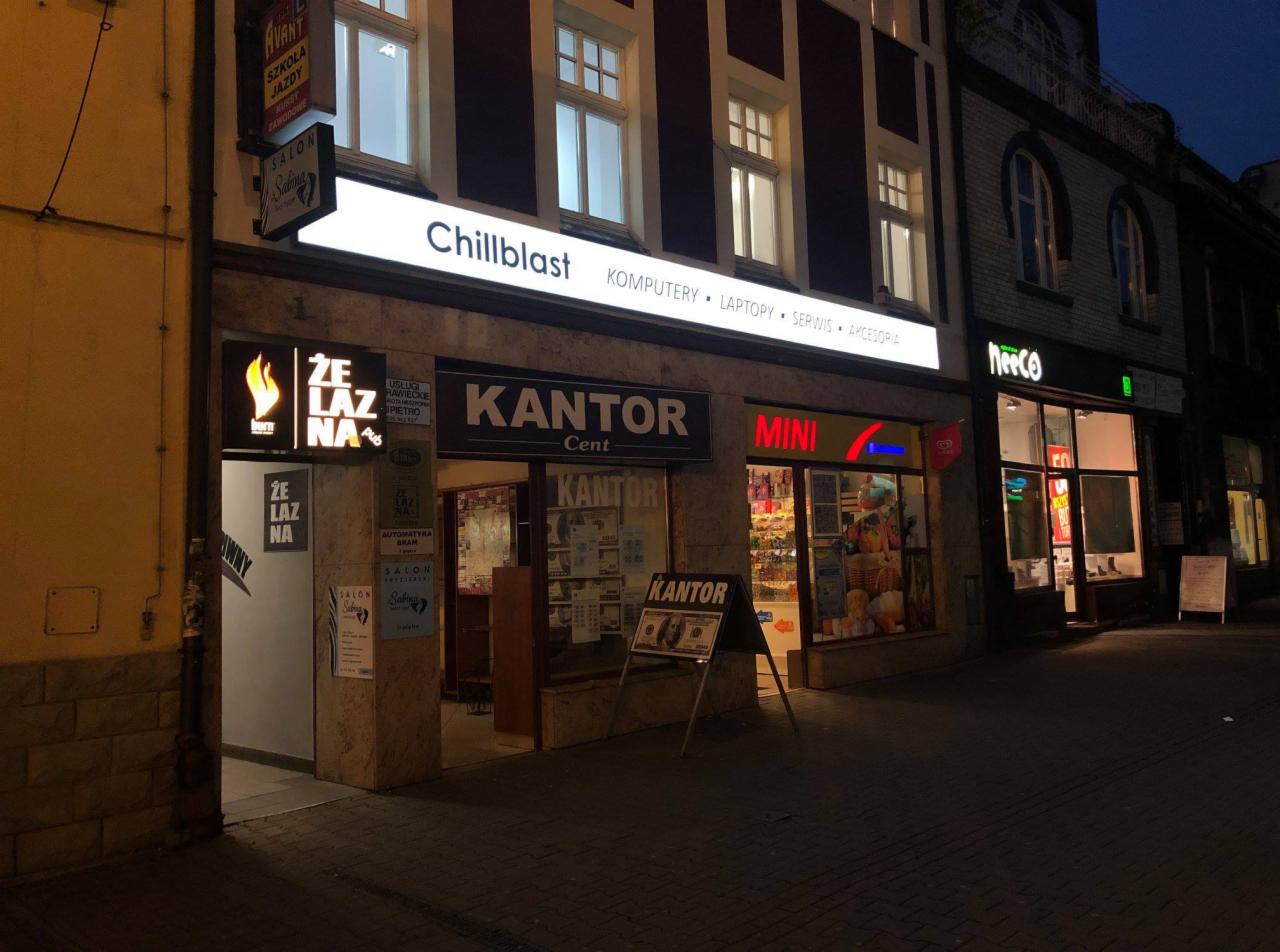 Fasada budynku z szyldem 'Chillblast' oferującego komputery, laptopy, serwis i akcesoria, widoczna wieczorem obok innych lokali usługowych.