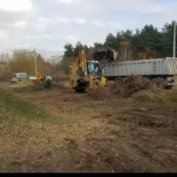 Żółta koparko-ładowarka z łyżką wsypuje rozdrobnione gałęzie do skrzyni ciężarówki na tle drzew i pochmurnego nieba, obok stosy zrębków i pracownicy przy rębaku.