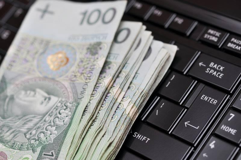 Plik banknotów 100 PLN leżący na czarnej klawiaturze laptopa, widoczne klawisze Enter, Backspace i Shift.