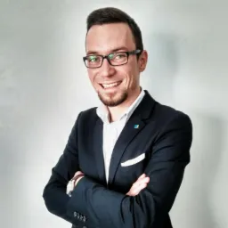 Uśmiechnięty mężczyzna w okularach i granatowym garniturze z białą poszetką, stoi z założonymi rękami na tle jasnoszarej ściany. Na klapie garnituru mała przypinka.