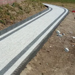 Zakrzywiony chodnik z jasnej kostki brukowej z ciemnymi pasami i obrzeżami, widoczne niedokończone prace i rozsypane kostki obok.