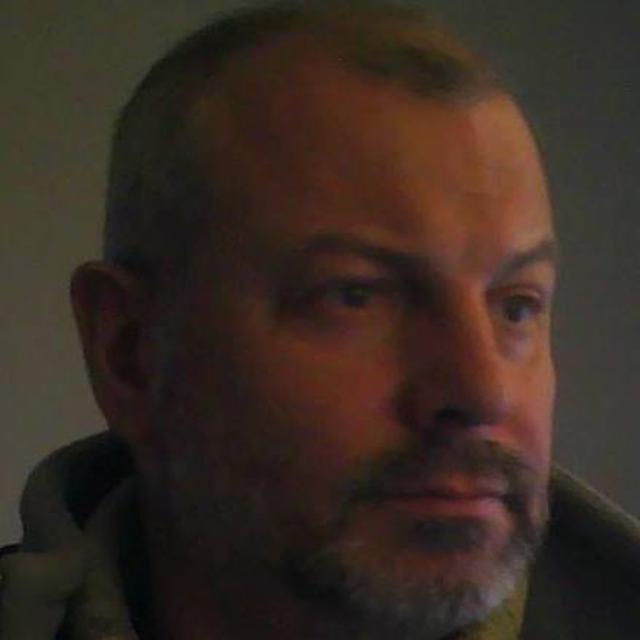 Portret mężczyzny z krótkimi włosami i zarostem, w ciemnym ubraniu, na rozmytym tle. Ujęcie z bliska, skupiające się na twarzy i wyrazie.