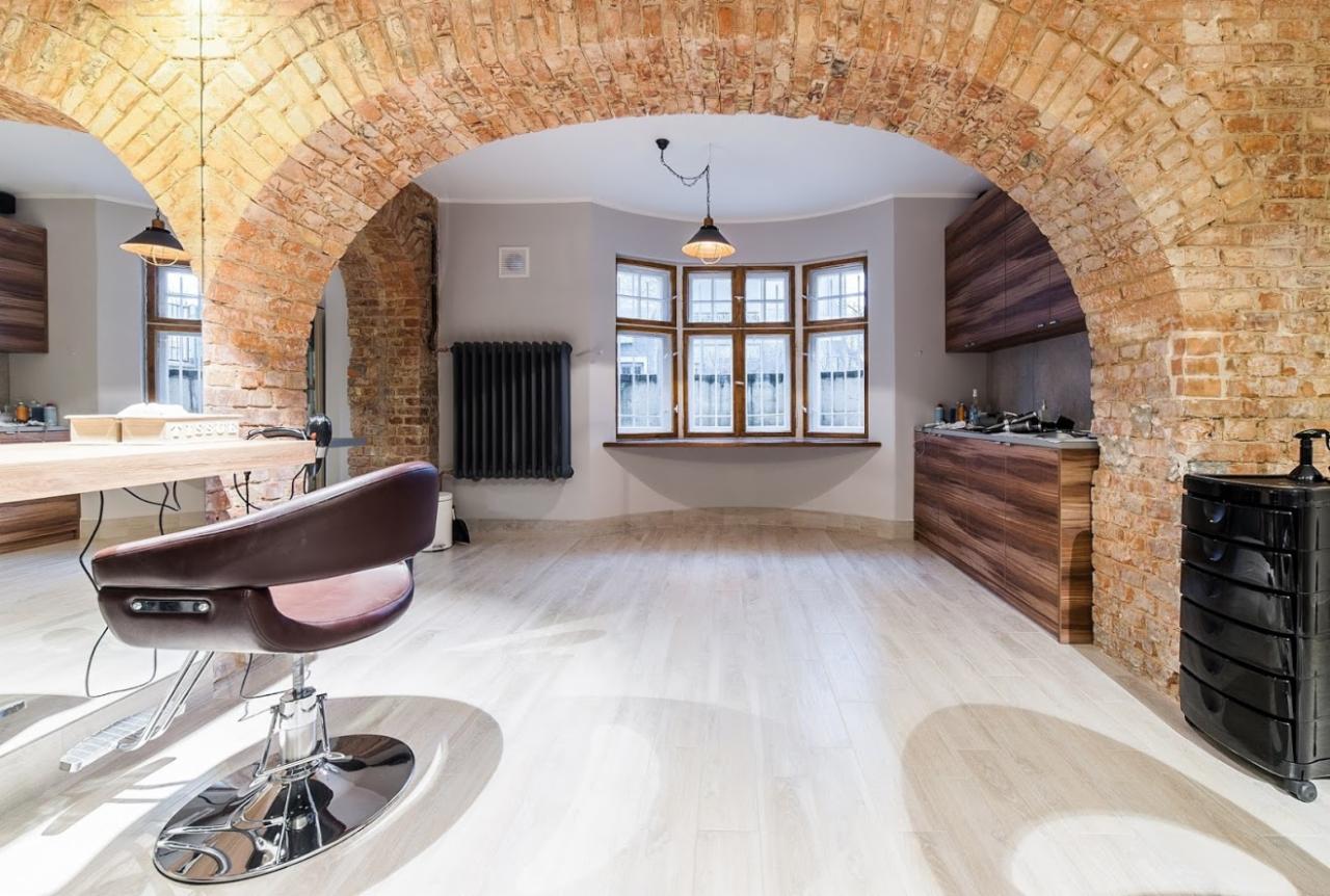 Przestronne wnętrze z ceglanym łukiem w Poznaniu, ukazujące zabudowę kuchenną z drewnianymi frontami i krzesło fryzjerskie, oświetlone wiszącą lampą.