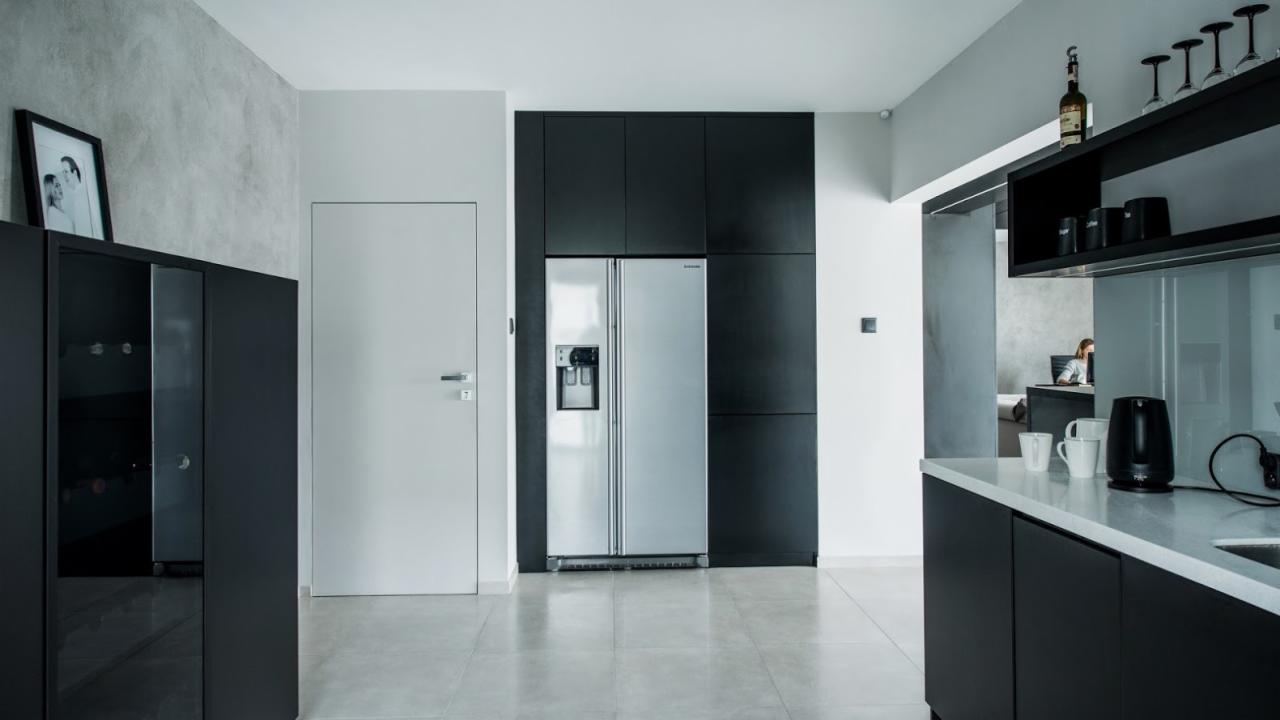 Nowoczesna kuchnia w odcieniach szarości i czerni z lodówką side-by-side w zabudowie, widok na blat z czajnikiem i filiżankami, w tle osoba pracująca przy biurku.