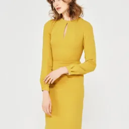 Kobieta prezentuje ołówkową sukienkę w odcieniu musztardowym z długimi rękawami i subtelnym wycięciem w dekolcie na białym tle.